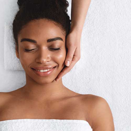 A woman getting a massage. She is laying on her back on a massage table. A therapist is placing their hand on her face.